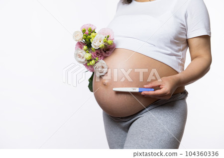 Close-up gravid woman, expectant mother holding a positive pregnancy inkjet test kit and bunch of spring flowers, above her naked pregnant belly, isolated white background. Copy space. Pregnancy time 104360336