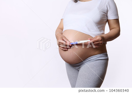 Close-up gravid woman, expectant mother holding a pregnancy inkjet test kit above her pregnant belly, isolated on white background. Fertility. Women's health. Child expectation. Pregnancy. Maternity 104360340