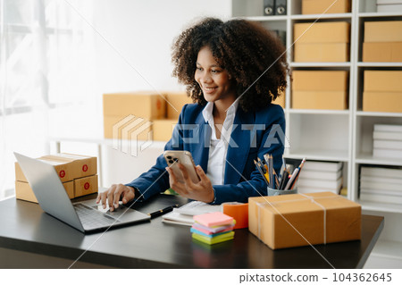 Startup small business SME, Entrepreneur owner African woman using smartphone or tablet taking receive and checking online purchase shopping order to preparing pack product box. . 104362645