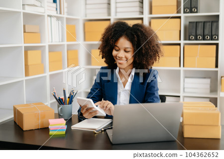 Startup small business SME, Entrepreneur owner African woman using smartphone or tablet taking receive and checking online purchase shopping order to preparing pack product box. . 104362652
