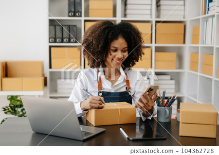 Startup small business SME, Entrepreneur owner African woman using smartphone or tablet taking receive and checking online purchase shopping order to preparing pack product box. . 104362728