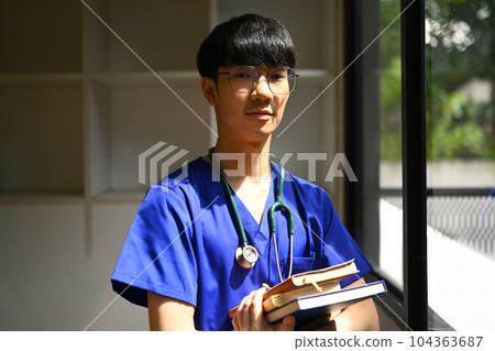 Young male medical students wearing blue scrubs with stethoscope standing near window in sunny day in medical office Young male medical students wearing blue scrubs with stethoscope standing near window in sunny day in medical office 104363687
