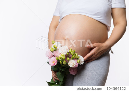Beauty close-up portrait of gravid expectant mother holding bouquet of flowers near her pregnant belly, gently caressing her abdomen, isolated white background. Women's health and gynecology concept Beauty close-up portrait of gravid expectant mother holding bouquet of flowers near her pregnant belly, gently caressing her abdomen, isolated white background. Women's health and gynecology concept 104364122