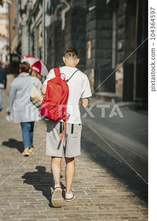 Young studen guy with backpack walking in city, holding smartphone and listening music headphones, rear view Young studen guy with backpack walking in city, holding smartphone and listening music headphones, rear view 104364597