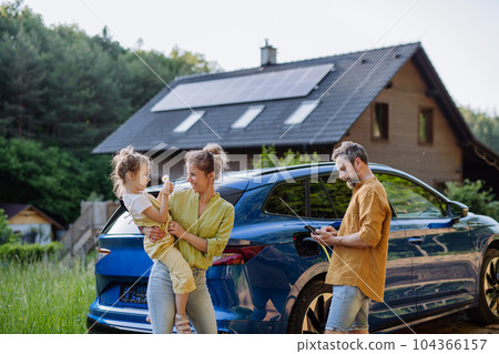 Family with little girl standing in front of their house with solar panels on the roof, having electric car. 104366157
