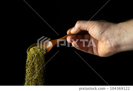 Mixture of dry chopped herbs is poured from a wooden spoon to add to a medicinal drink or tea. Free space for advertising on a black background. 104369122