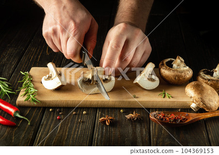 Close-up of a chef hands with a knife cutting mushrooms on a cutting kitchen board while preparing a vegetarian breakfast. Delicious mushroom diet with spices 104369153