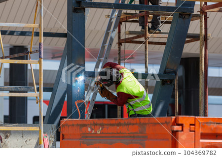 A man-welder fastens iron girders with a welding machine. Construction of the station. A man-welder fastens iron girders with a welding machine. Construction of the station. 104369782