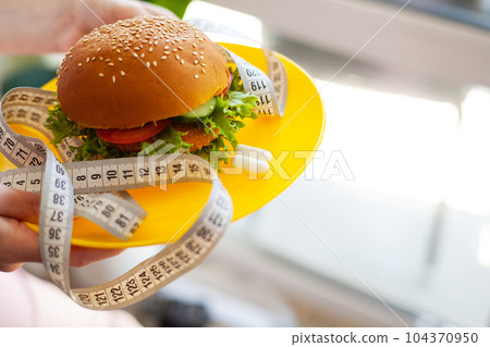 Woman holding unhealthy burger and tape measure, weight loss concept 104370950