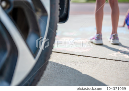 Selective focus on pavement with a vehicle tire in the foreground and a blurred child standing in the path.  104371069