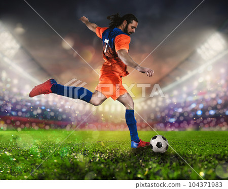 Soccer scene at night match with player in a orange uniform kicking the ball with power 104371983