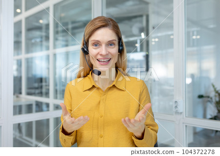 Mature adult business woman with blond hair working inside light office, customer service tech worker smiling and talking to customer consulting, using headset and laptop for remote video call. 104372278