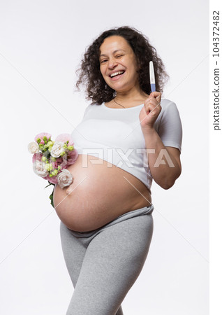 Smiling happy pregnant woman showing at camera a positive pregnancy test, posing with bunch of flowers, isolated over white background. Human fertility. Gynecology. Female health and maternity concept Smiling happy pregnant woman showing at camera a positive pregnancy test, posing with bunch of flowers, isolated over white background. Human fertility. Gynecology. Female health and maternity concept 104372482