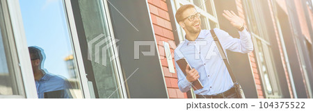 Young happy caucasian businessman wearing blue shirt and eyeglasses holding his laptop and waving, saying hello while standing against office building outdoors Young happy caucasian businessman wearing blue shirt and eyeglasses holding his laptop and waving, saying hello while standing against office building outdoors 104375522