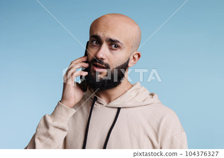 Unsure arab man talking on smartphone and looking away with uncertain expression. Young bald bearded person in doubt while answering mobile phone call and having communication Unsure arab man talking on smartphone and looking away with uncertain expression. Young bald bearded person in doubt while answering mobile phone call and having communication 104376327