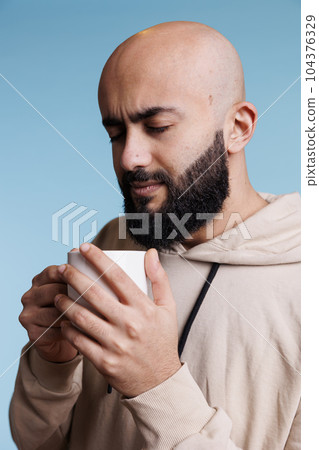 Relaxed arab man holding white cup in hands while enjoying coffee. Young thoughtful person wearing casual hoodie clothes drinking hot tea beverage from mug on blue background 104376329