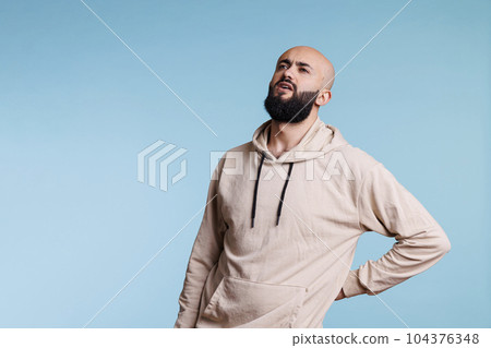 Young arab man holding hand on lower back while having spine problems. Bearded person looking upwards in pain and grimacing while suffering from backache and sore muscles cramps Young arab man holding hand on lower back while having spine problems. Bearded person looking upwards in pain and grimacing while suffering from backache and sore muscles cramps 104376348