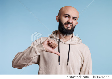 Arab man showing dislike with thumb down, expressing negative emotion portrait. Young person with disapprovement face grimace posing with disagreement sign and looking at camera Arab man showing dislike with thumb down, expressing negative emotion portrait. Young person with disapprovement face grimace posing with disagreement sign and looking at camera 104376366