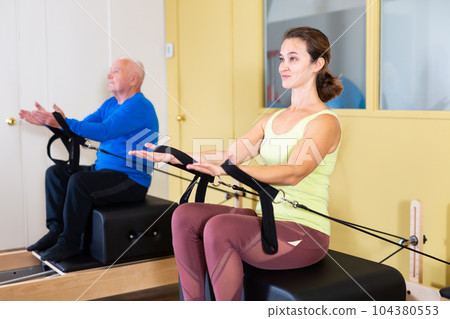 Woman exercising on Pilates reformer with elderly man in rehab 104380553