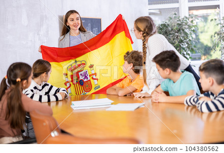 Smiling young female teacher explaining culture of country while showing Spanish flag to schoolchildren preteens in classroom 104380562