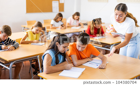 Friendly teacher woman helping children during lesson in schoolroom Friendly teacher woman helping children during lesson in schoolroom 104380704