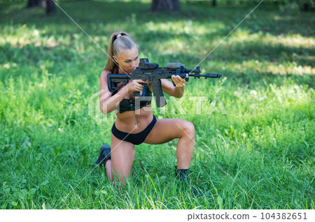 Caucasian woman in bikini sits with machine gun on green grass. Caucasian woman in bikini sits with machine gun on green grass. 104382651