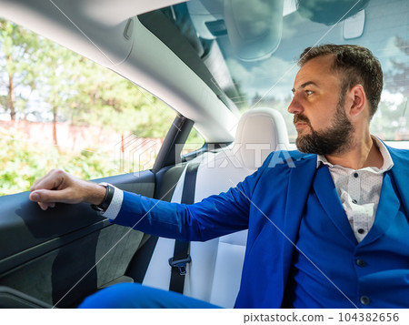 A caucasian man in a blue suit looks out the open window while sitting in the back seat of a car. Business class passenger. A caucasian man in a blue suit looks out the open window while sitting in the back seat of a car. Business class passenger. 104382656