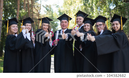 A group of graduates in robes give a thumbs up outdoors. Elderly student. 104382682