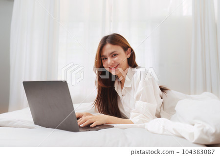 Happy smiling asian woman stretching on bed in the morning and using laptop computer 104383087