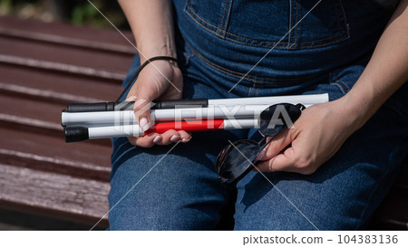 A blind woman sits on a bench and holds a folding tactile cane and sunglasses in her hands. A blind woman sits on a bench and holds a folding tactile cane and sunglasses in her hands. 104383136