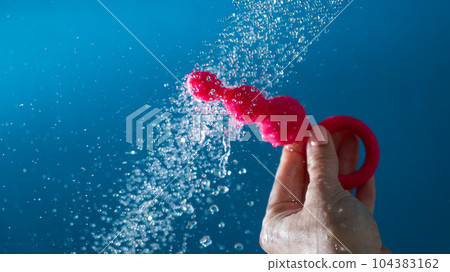 Woman holding pink anal beads under running water on blue background. Sex toy hygiene concept. Woman holding pink anal beads under running water on blue background. Sex toy hygiene concept. 104383162