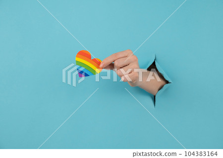 A hand with a rainbow-colored heart sticking out of a hole in a blue cardboard background.  104383164