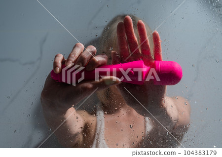 A woman in a white tank top stands in the shower and holds a pink sex toy. A woman in a white tank top stands in the shower and holds a pink sex toy. 104383179