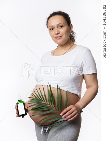 Gorgeous multi ethnic pregnant woman, looking at camera, holding exotic palm leaf cosmetic oil massage over her big belly, isolated white background. Skin and body care at pregnancy maternity time Gorgeous multi ethnic pregnant woman, looking at camera, holding exotic palm leaf cosmetic oil massage over her big belly, isolated white background. Skin and body care at pregnancy maternity time 104386382