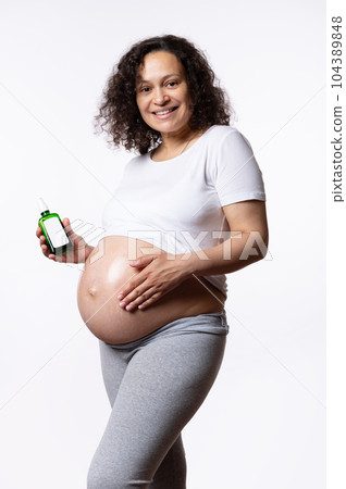 Multi ethnic beautiful pregnant woman smiles looking at camera, holds a mockup jar with skin care product, applying stretch marks cosmetic oil, massaging her big belly over white isolated background. Multi ethnic beautiful pregnant woman smiles looking at camera, holds a mockup jar with skin care product, applying stretch marks cosmetic oil, massaging her big belly over white isolated background. 104389848