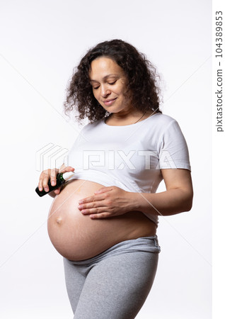 Vertical advertising studio portrait of expectant pregnant woman applying massage oil, moisturizing her big belly to avoid stretch marks during childbearing and pregnancy time, isolated white backdrop 104389853