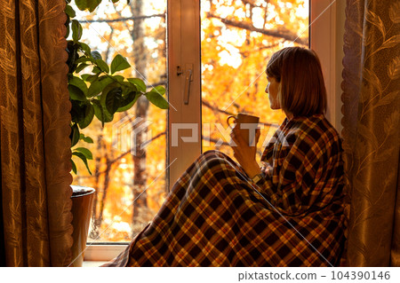 Autumn coffee. A beautiful girl with a cup of coffee in her hands sits and looks out the window. Cozy autumn composition. Copy space 104390146