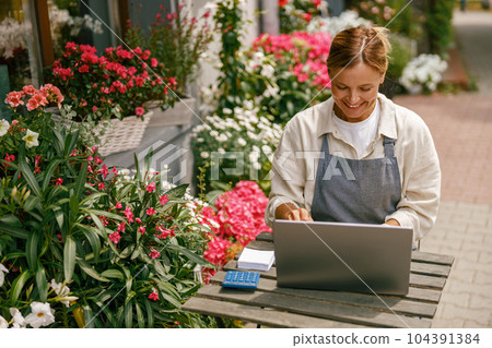 Smiling florist working on laptop and taking online orders in store Smiling florist working on laptop and taking online orders in store 104391384