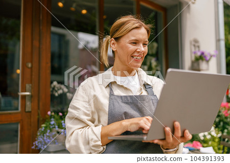Small flower shop owner florist wearing apron working on laptop and taking online orders in store Small flower shop owner florist wearing apron working on laptop and taking online orders in store 104391393