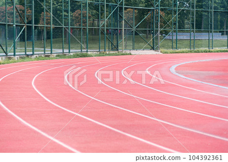 All-weather running track with red rubberized surface separated by white marking lines for athletics training at contemporary sports ground in urban park on summer day All-weather running track with red rubberized surface separated by white marking lines for athletics training at contemporary sports ground in urban park on summer day 104392361