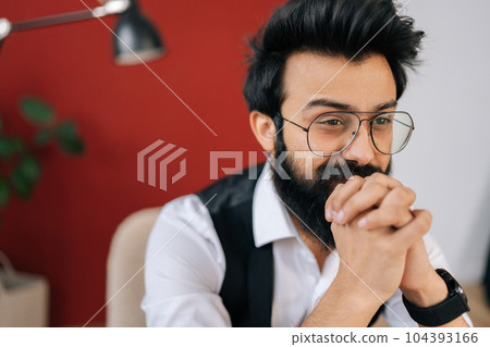 Close-up face of handsome Indian businessman wearing elegant formalwear sitting on chair and smiling looking away. Front view of friendly Indian young man in fashion glasses posing at studio. Close-up face of handsome Indian businessman wearing elegant formalwear sitting on chair and smiling looking away. Front view of friendly Indian young man in fashion glasses posing at studio. 104393166