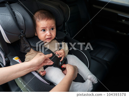 mother is fastening safety belt to crying infant baby in car seat 104393378