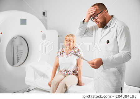 Medical computed tomography or MRI scanner. Shocked, surprised doctor looking at scan, examining results of MRI. Female patient sitting on couch on blurred background. Concept of healthcare. 104394195