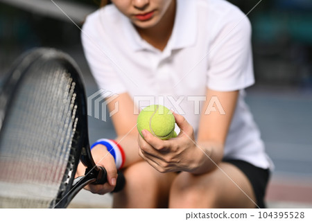 Pensive young female tennis player holding ball racket sitting on the bench at tennis court 104395528