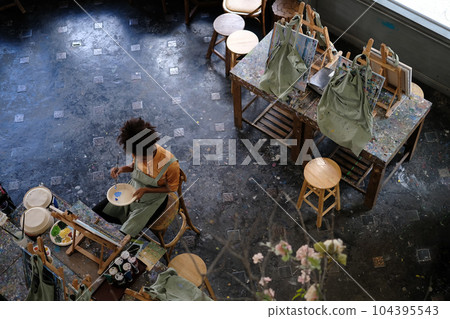 Overhead view of young African American female student painting picture on canvas, sitting alone in art workshop Overhead view of young African American female student painting picture on canvas, sitting alone in art workshop 104395543