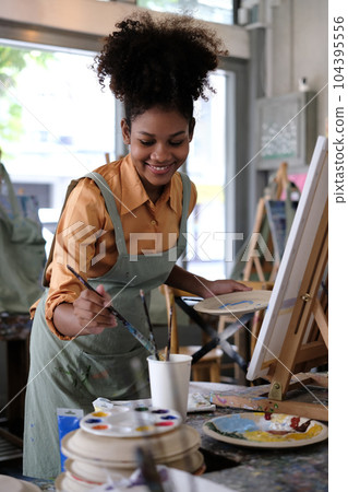 Happy young curly art school female student painting picture with brush on canvas in art studio Happy young curly art school female student painting picture with brush on canvas in art studio 104395556