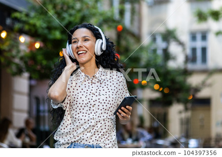 Beautiful young Latin American woman walks in the evening city on a trip, woman with headphones listens to music and dances and sings, uses an online radio application on her phone 104397356