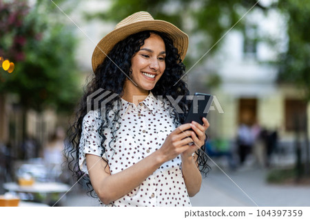 A beautiful young woman walks through the evening city in a hat, a smiling Latin American woman holds a smartphone in her hands. A tourist with curly hair types a message and browses online pages on A beautiful young woman walks through the evening city in a hat, a smiling Latin American woman holds a smartphone in her hands. A tourist with curly hair types a message and browses online pages on 104397359