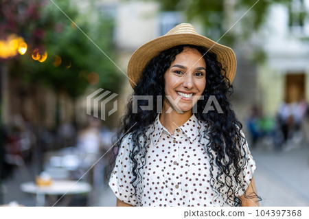 Beautiful young Latin American woman portrait, woman walking in evening city in hat with curly hair in warm weather, smiling and looking at camera close up. Beautiful young Latin American woman portrait, woman walking in evening city in hat with curly hair in warm weather, smiling and looking at camera close up. 104397368