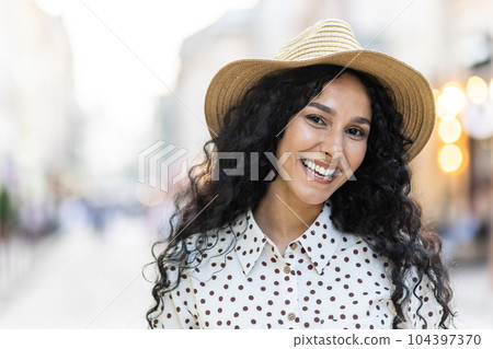 Beautiful young Latin American woman portrait, woman walking in evening city in hat with curly hair in warm weather, smiling and looking at camera close up. Beautiful young Latin American woman portrait, woman walking in evening city in hat with curly hair in warm weather, smiling and looking at camera close up. 104397370